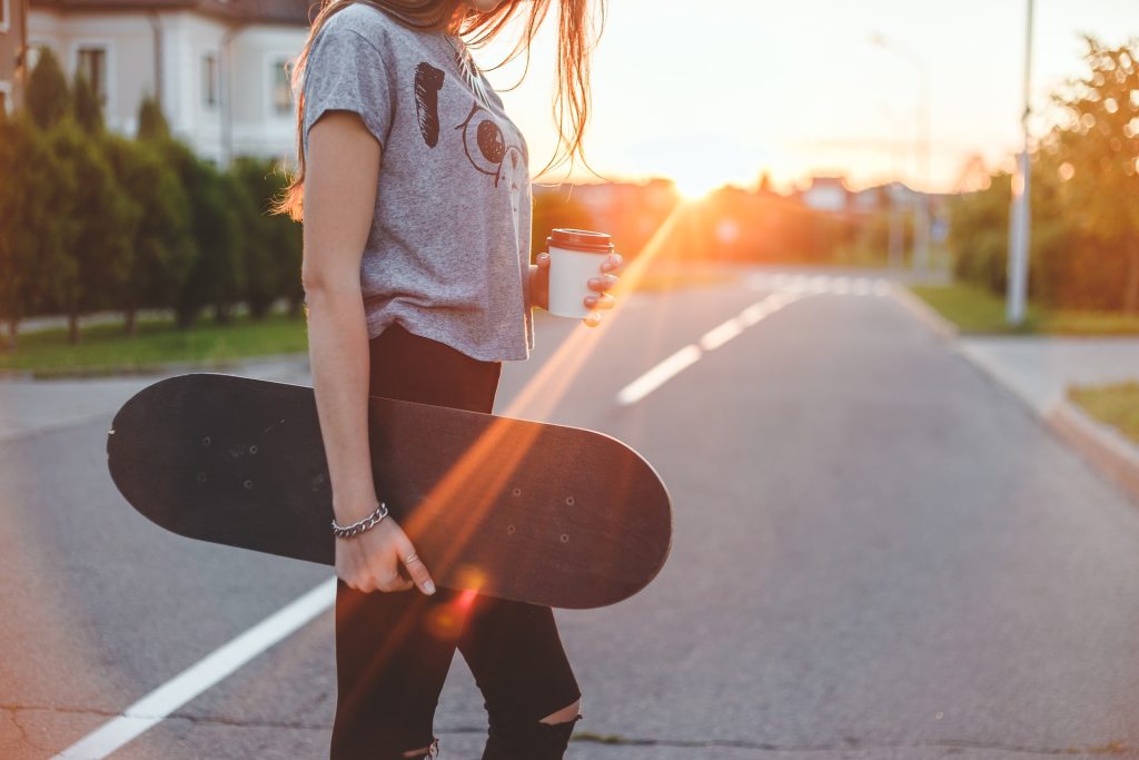 Mädchen mit Skateboard und Coffee-to-go-Becher im Sonnenuntergang